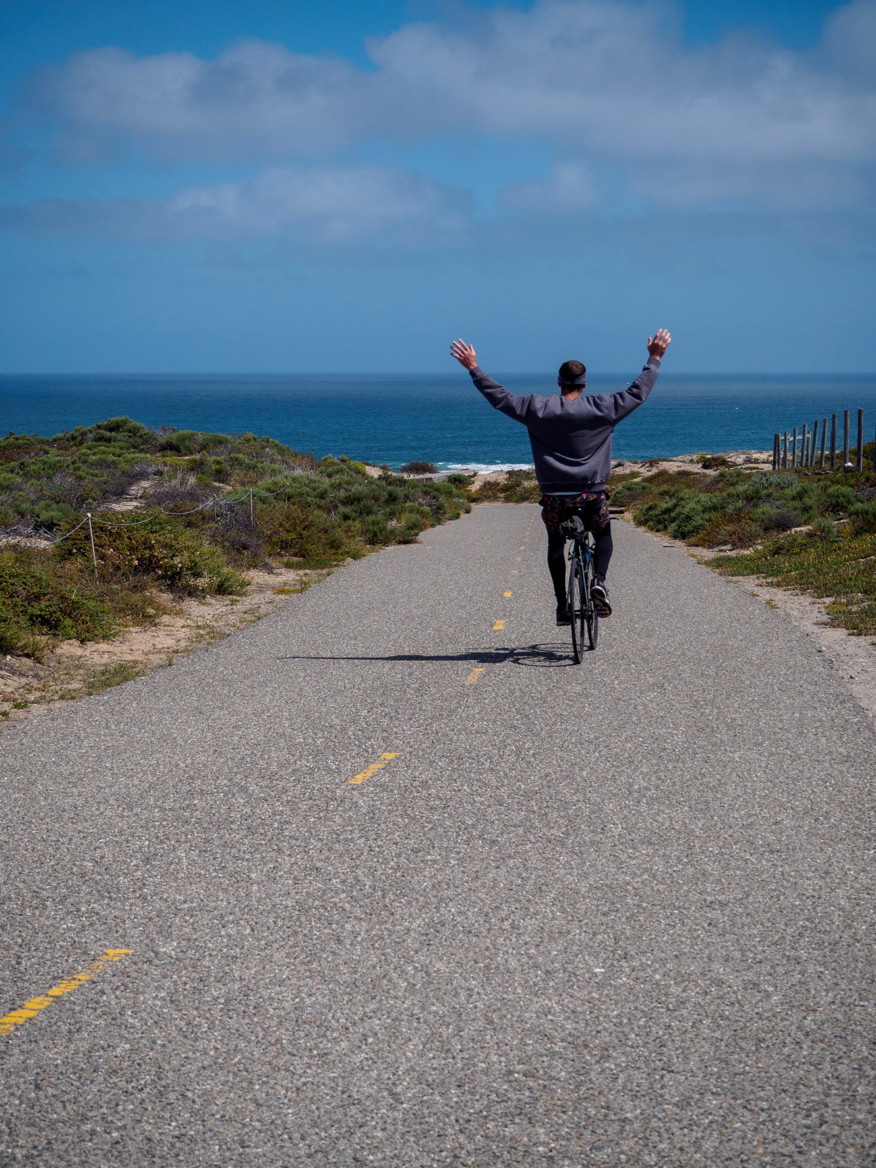 A cyclist riding down a paved hill towards the beach with his arms up in the air