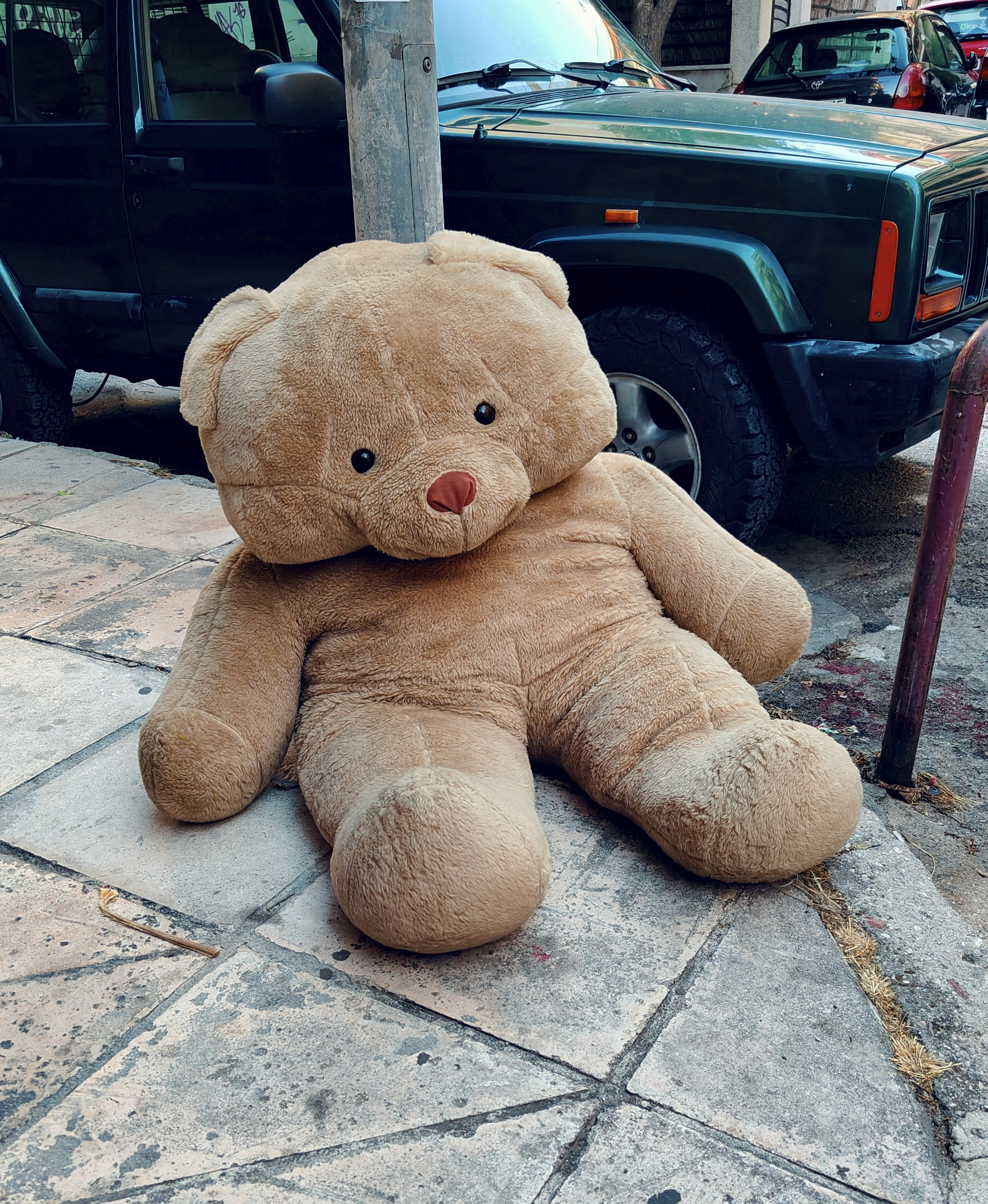 A gigantic brown teddy bear left leaning against a pole on the corner of a sidewalk.